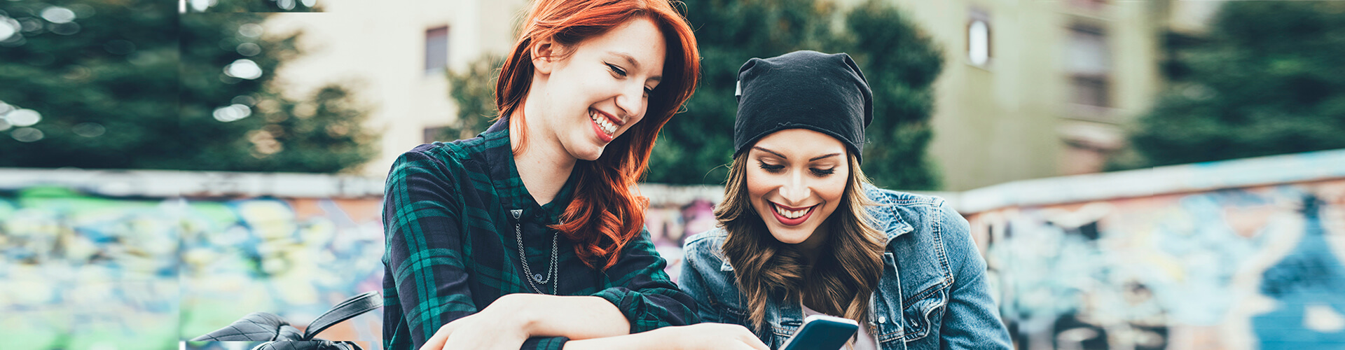 Programa Joven In - Dos chicas jóvenes sonriendo mientras están con el móvil en la mano sentadas en un parque Programa Joven In - Dos chicas jóvenes sonriendo mientras están con el móvil en la mano sentadas en un parque
