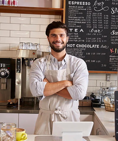 Ventajas para Autonomos - El dueño de una cafeteria  con los brazo cruzados sonriendo tras la barra de su negocio Ventajas para Autonomos - El dueño de una cafeteria  con los brazo cruzados sonriendo tras la barra de su negocio