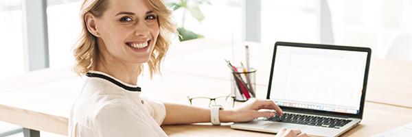 Lineas ICO - Mujer joven rubia de negocios sonriendo mientras se encuentra sentada en su escritorio con su portatil